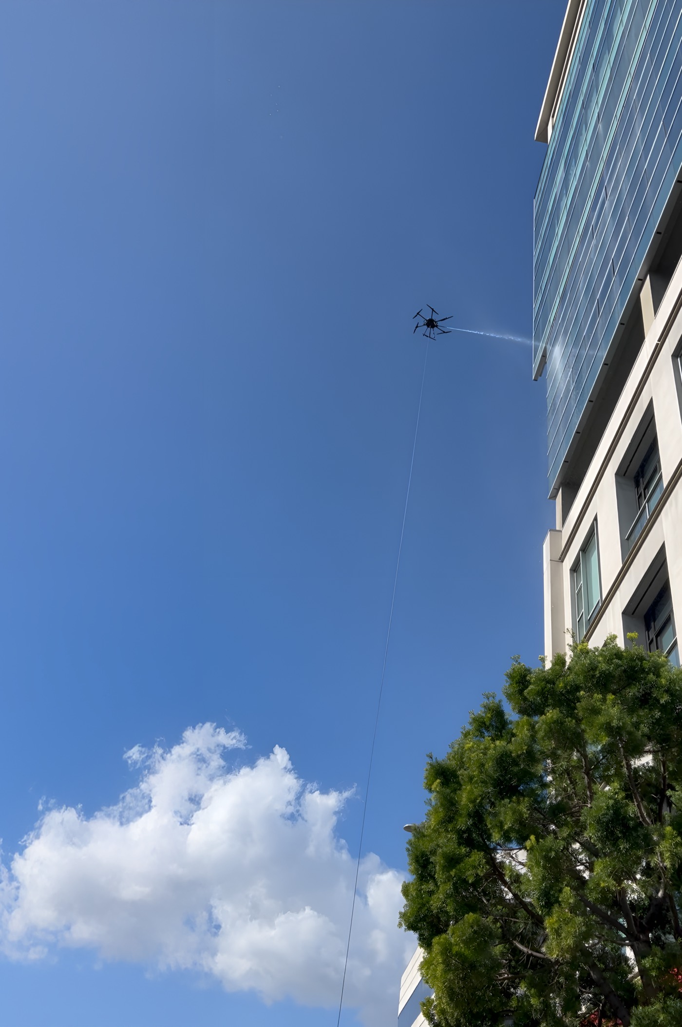 A Stratos drone spraying the glass facade of an office building, tethered to the ground crew below.